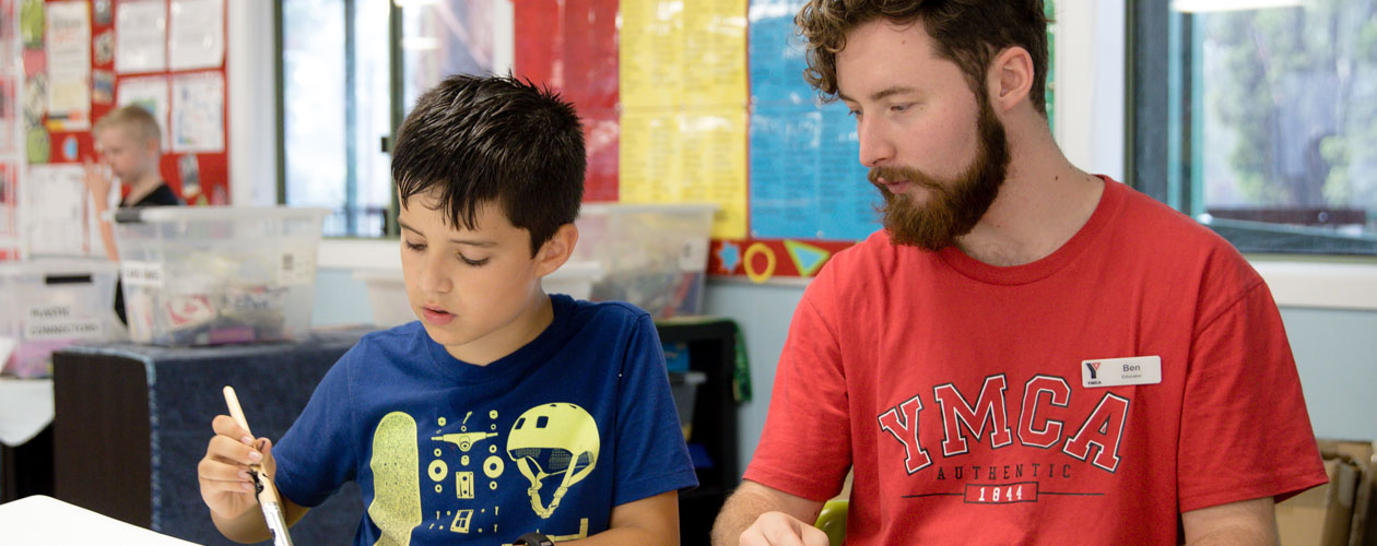 YMCA staff member sitting next to a young boy with a paintbrush