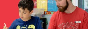 YMCA staff member sitting next to a young boy with a paintbrush