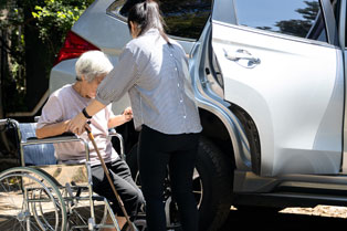 aged care worker helping an older woman out of a wheelchair and into a car