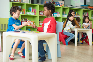 2 children sitting at table in preschool setting includes a teacher and additional students at another table in background