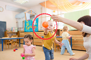A childcare worker holds a hoop while a small boy throws a ball, with other children playing nearby