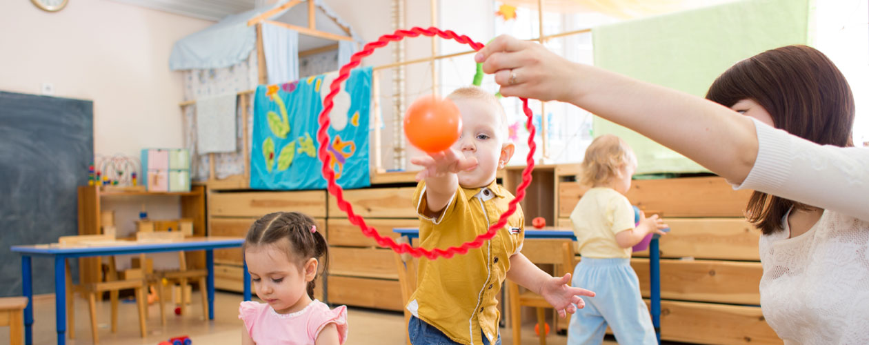 A childcare worker holds a hoop while a small boy throws a ball, with other children playing nearby