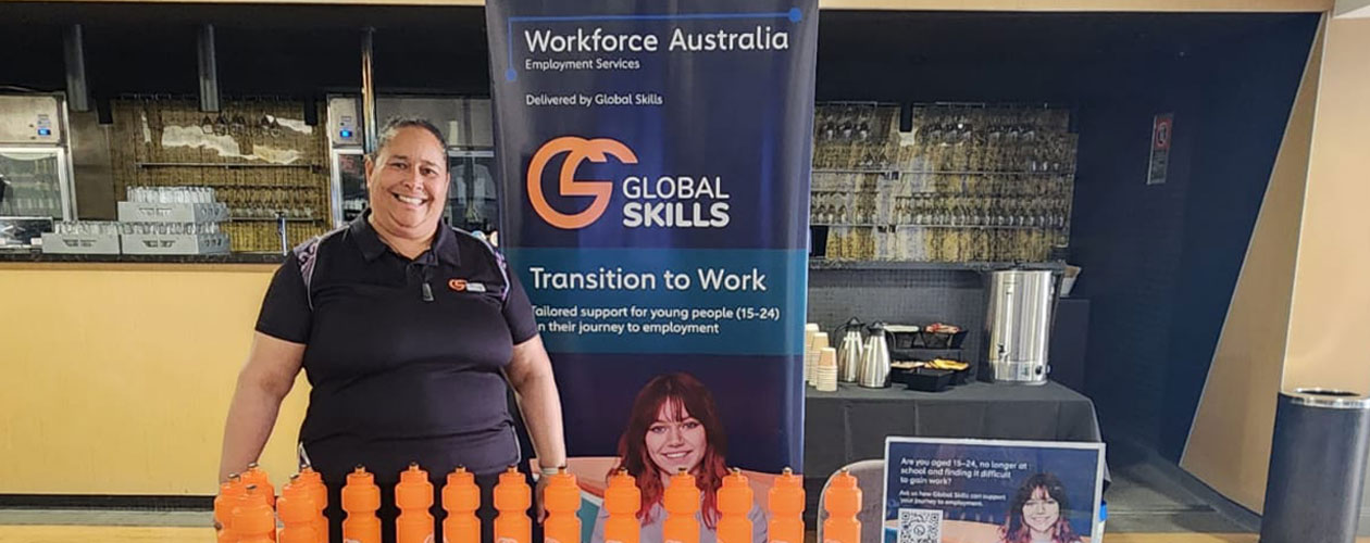 Jennifer standing behind our expo table, next to a Global Skills Transition to Work banner, with branded bottles and merchandise on display.