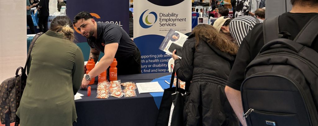 Youssef engaging with expo attendees. Our stall features banners for Workforce Australia and Disability Employment Services, with promotional items like orange water bottles and brochures on the table.