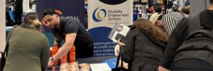 Youssef engaging with expo attendees. Our stall features banners for Workforce Australia and Disability Employment Services, with promotional items like orange water bottles and brochures on the table.