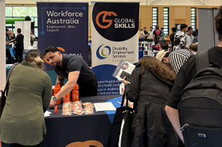 Youssef engaging with expo attendees. Our stall features banners for Workforce Australia and Disability Employment Services, with promotional items like orange water bottles and brochures on the table.