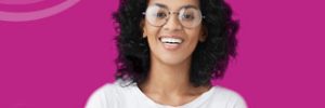 dark, curly haired woman wearing a white shirt and glass standing again a pink background