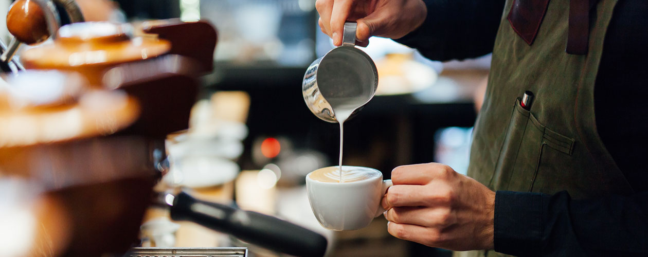 A close-up of a person pouring steamed milk from a metal jug into a white coffee cup. The background is slightly blurred, with coffee equipment visible.