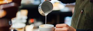 A close-up of a person pouring steamed milk from a metal jug into a white coffee cup. The background is slightly blurred, with coffee equipment visible.