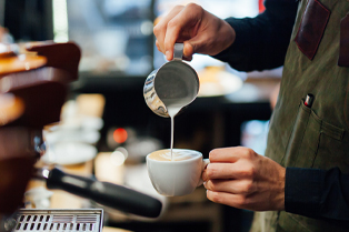 A close-up of a person pouring steamed milk from a metal jug into a white coffee cup. The background is slightly blurred, with coffee equipment visible.