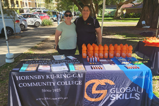 Jennifer and Zeina standing behind our shared stall at the event. The table is covered with branded tablecloths and displays brochures, pens, water bottles, and other promotional items.