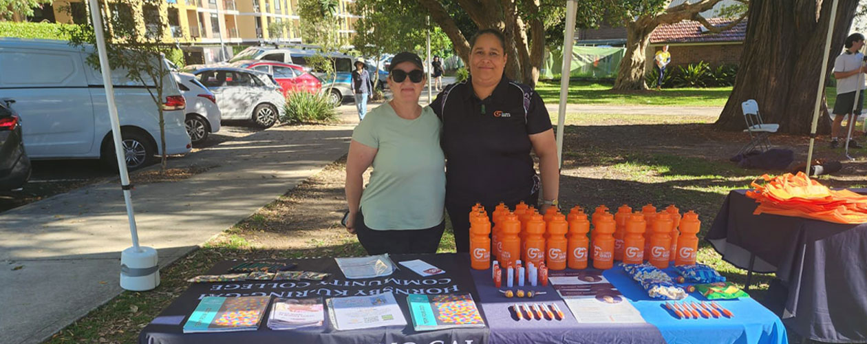 Jennifer and Zeina standing behind our shared stall at the event. The table is covered with branded tablecloths and displays brochures, pens, water bottles, and other promotional items.
