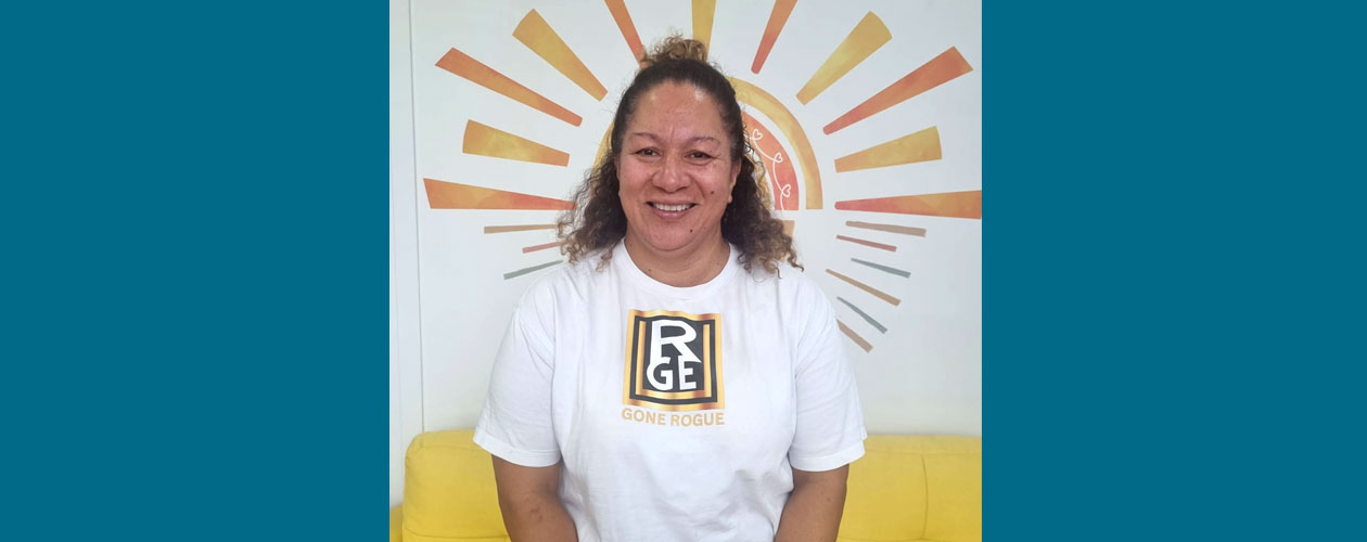 Ashley standing in front of a painted sunrise wall, wearing a white “Gone Rogue” t-shirt.