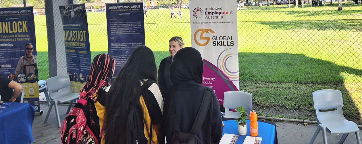Jessica is engaging with students at the Global Skills stall during the expo. A Global Skills and Inclusive Employment Australia banner is behind her, with a sports field in the background.