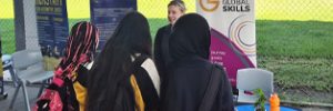 Jessica is engaging with students at the Global Skills stall during the expo. A Global Skills and Inclusive Employment Australia banner is behind her, with a sports field in the background.