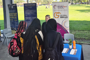 Jessica is engaging with students at the Global Skills stall during the expo. A Global Skills and Inclusive Employment Australia banner is behind her, with a sports field in the background.