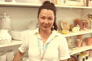Liisa at work standing beside shelves displaying homewares such as vases, bowls and plates. At the bottom of the image is a teal banner with the Workforce Australia and Global Skills logos.