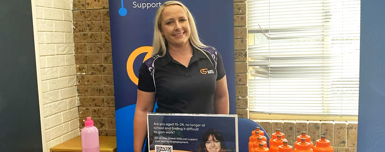 ess standing at Global Skills stall in front of a Workforce Australia banner. The stall is set up with brochures, pens, snacks and branded water bottles, along with a sign featuring a QR code for more information.
