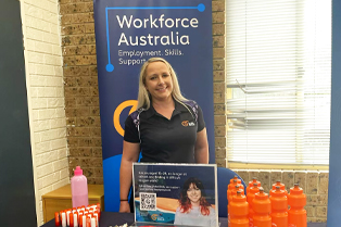ess standing at Global Skills stall in front of a Workforce Australia banner. The stall is set up with brochures, pens, snacks and branded water bottles, along with a sign featuring a QR code for more information.