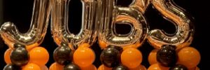 Large metallic “JOBS” balloon letters are surrounded by orange, black, and gold balloons.