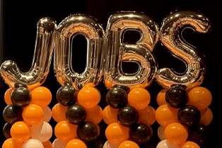 Large metallic “JOBS” balloon letters are surrounded by orange, black, and gold balloons.