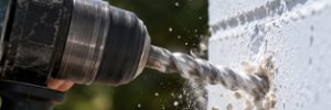 A person drills into a white brick wall outdoors, with masonry dust scattering.