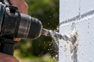 A person drills into a white brick wall outdoors, with masonry dust scattering.