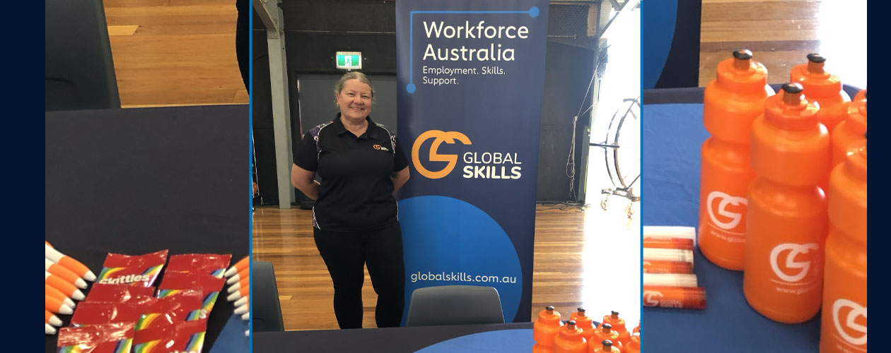 Global Skills team member, Melinda, standing beside a Workforce Australia banner at the event, with a promotional table displaying branded pens, drink bottles and other materials.