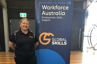 Global Skills team member, Melinda, standing beside a Workforce Australia banner at the event, with a promotional table displaying branded pens, drink bottles and other materials.