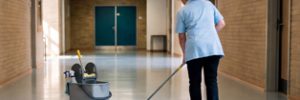 A cleaner in a blue uniform mops the floor of a school corridor, with a cleaning trolley beside her.